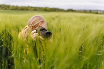 Female nature photographer with blond hair taking photos of green wheat field during spring windy...