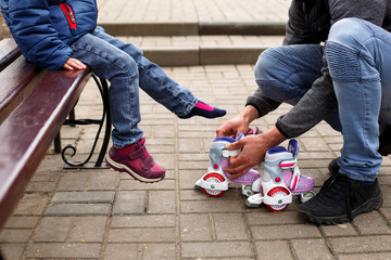 Dad helps his little daughter put on roller skates. Parental care, father's day, family weekend.