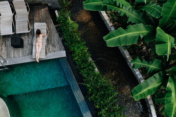 Fototapeta na wymiar Top view of unrecognizable slim young woman in beige bikini relaxing and sunbathe near luxury swimming pool in green tropic in Ubud.Vacation concept. Drone photo
