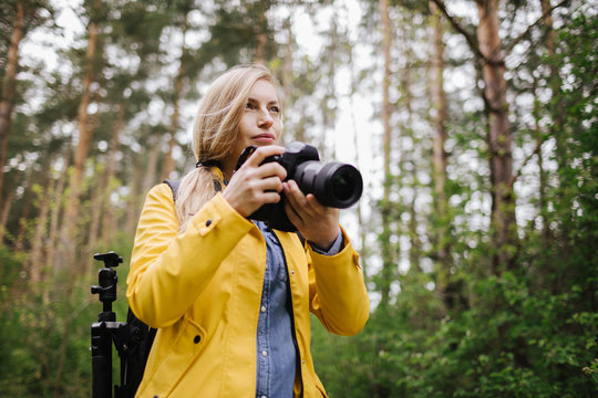 Concentrated woman in yellow jacket using professional camera for taking pictures of beautiful wild nature at green forest. Concept of photography and adventure.