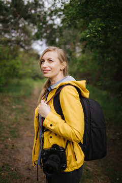Charming mature woman with blond hair standing among spring forest with big backpack and professional camera. Photographer in yellow jacket working on fresh air