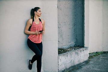 Young attractive slim sportswoman leaning on the wall and holding smart phone while resting after exercising.