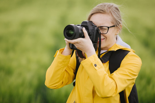 Portrait of charming female photographer with blond hair wearing eyeglasses and yellow jacket while taking pictures of beautiful nature. Blur background of green wheat field.