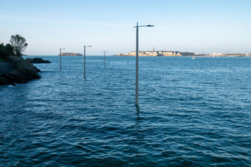 Dinard, France 24-05-2020. Ocean tide view of the flooded port road in the distance the cities of Saint Malo and Intramuros. Street lanterns protruding into the water - obrazy, fototapety, plakaty