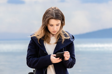 A young adult girl with a smartphone stands on the seashore against a dark cloudy sky.