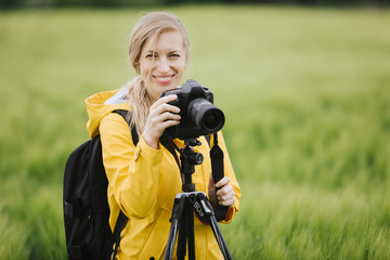 Portrait of attractive woman in eyeglasses and yellow jacket standing among wheat field with tripod and digital camera. Smiling female photographer enjoying spring weather outdoors. - obrazy, fototapety, plakaty