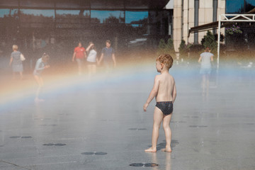 little boy with blond hair walking around the city and enjoying the sunshine. Fountain with water making a rainbow.