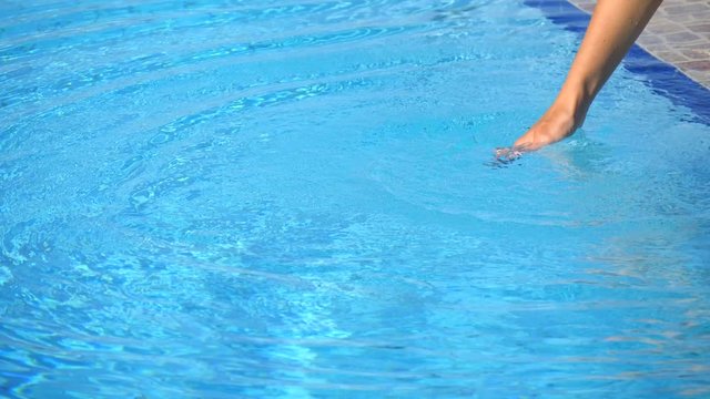 Female hand of young girl tests water in pool, touches water, pours water from hand, hits the water with his hand, creates spray, circles on water. Summer holidays, relaxation, spa, vacation, fun.