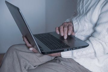 A man in a white sweater sits on a chair, holds a laptop in his hands and works. Only hands and a laptop are visible. The background is white.