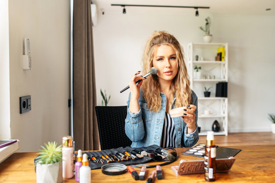 A young well-groomed woman with a smooth skin doing self makeup at home, she sits at the desk and looks at mirror, cosmetics products on the table