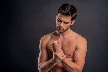 Fototapeta premium Handsome young bearded man isolated. Cropped image of topless muscular man is standing on gray background. Man holding his wrist. Experiencing wrist pain.