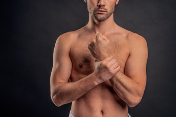 Handsome young bearded man isolated. Cropped image of topless muscular man is standing on gray background. Close-up of man holding his wrist. Experiencing wrist pain.