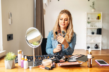 Female professional makeup artist, a makeup teacher with a different brushes in hands shows their into camera. Cosmetics products and mirror on the table