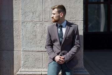 Portrait of a young bearded guy of twenty-five years old, in a business suit, against the background of an office building. Posing looking at the camera. Outdoors