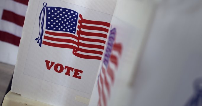 Close Up, Angled View Across Voting Booths At Polling Station In United States During Election. Large US Flag On Wall Behind.