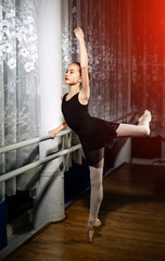 Obraz na płótnie Canvas Attractive ballerina with her hair in bunch stretcing leg near the window. Young girl in black costume, white tights and pointes training in a ballet studio.