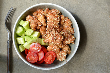 Tasty lunch in a gray bowl on a concrete background. Nuggets with sesame seeds. Cucumbers and tomatoes. Salad and chicken. Free space for text Top view