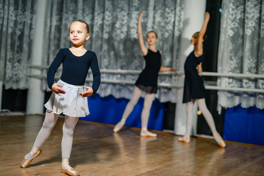 Little ballerina standing in pose with her hands on a blur background with two ballerinas. Girls practicing during the lesson in a dancing studio. Close-up