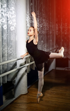 Attractive ballerina with her hair in bunch stretcing leg near the window. Young girl in black costume, white tights and pointes training in a ballet studio.