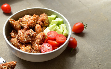 Tasty lunch in a gray bowl on a concrete background. Nuggets with sesame seeds. Cucumbers and tomatoes. Salad and chicken. Free space for text