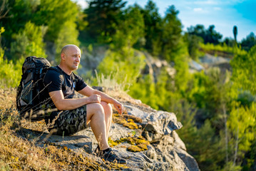 Traveler looks at the landscape around himself at the top of the cliff in the summer in warm weather. A man sits on squat near a cliff in a sunny day