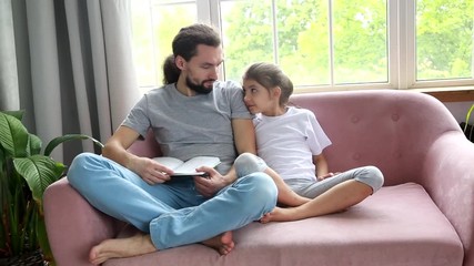 Father day and happy fatherhood concept.Close up father and daughter are reading a book at living room. Father hugs daughter and smiles. It is pleasant to be at home with family during bad weather.
