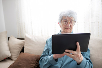 Sad and lonely senior gray-haired Caucasian woman sitting on a sofa in brightly lit living room and browsing internet or looking at family photos on a tablet