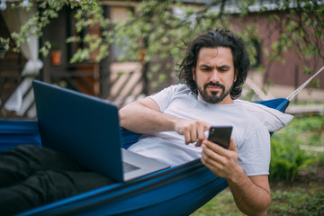 A man works with a laptop and a phone in a hammock in a country house