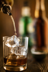 In the bar, whiskey is poured into a glass with ice, in the background there are bottles on a wooden table of the bar counter, shallow depth of field, selective focus. The concept of alcoholic drinks