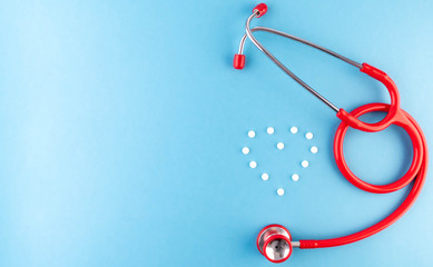 Medical equipment: red stethoscope and a heart shape of white pills on blue background with a place for text. Medical template or mockup. Cardiology concept.