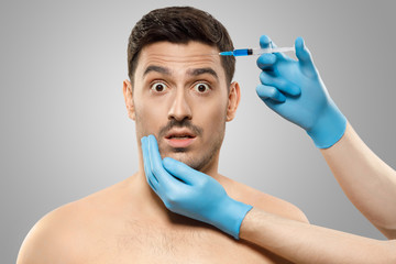 Young male afraid of beauty procedures, and plastic surgery, having rejuvenating injection in forehead by doctor in gloves, isolated on gray background