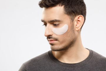 Obraz premium Young male looking aside and down with eye patch preventing from dark circles and wrinkles, taking care of his face skin, isolated on gray background