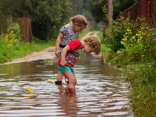 Young children in rubber boots with toys on a rope in a deep pool after a rain peer into the water.