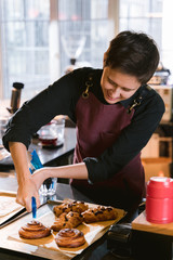 Woman chef baker barista in apron during process of glaze buns with caramel. Smiley female person with short brunette hair cut preparing pastry in an authentic coffee shop interior.