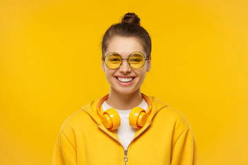 Young teenage girl wearing t-shirt, hoodie, round colored eyeglasses and wireless headphones around neck, laughing happily, isolated on yellow background