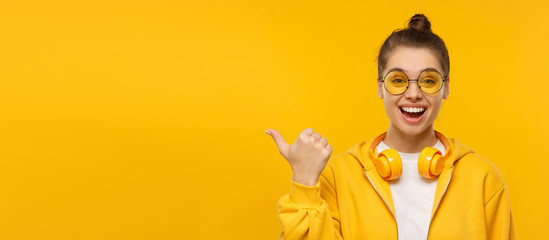 Horizontal banner of young excited female wearing t-shirt, hoodie, glasses and wireless headphones around neck, pointing to copy space, isolated on yellow background