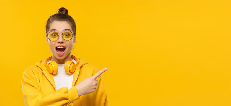 Horizontal banner of young shocked girl dressed in t-shirt and hoodie, wearing round glasses and headphones on neck, pointing to copy space, isolated on yellow background