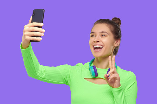 Young happy girl wearing green neon tep and headphones around neck, taking selfie photo with her phone, smiling and showing V sign, isolated on purple backgroound