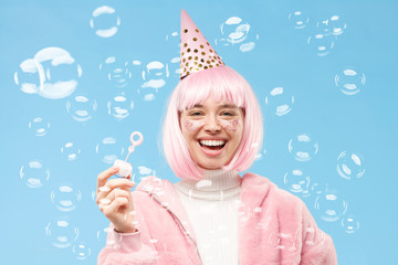 Young laughing teenage girl wearing pink wig, coat and hat for her birthday party, blowing soap...