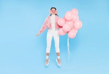 Full length photo of young happy teenage girl, wearing winter clothes and jumping with bunch of pink helium balloons, isolated on blue background