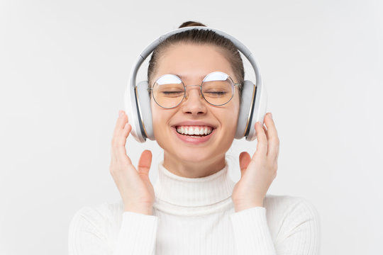 Young female with closed eyes, wearing glasses and wireless headphones, enjoying listening to music and smiling happily, isolated on gray background