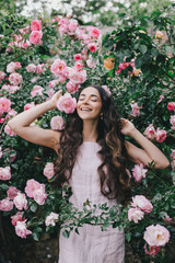 Obraz na płótnie Canvas Beautiful young woman with long curly hair and perfect skin wearing pink linen dress posing near blooming roses in a garden. Nude make up. Close up portrait