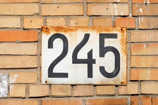 House number 245 (two hundred and forty five). Black lettering on a rusty white metal plate on an old brick wall