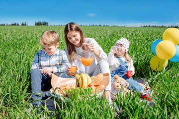 Three happy kids sitting on picnic on the field. blue sky, green grass. bread, pies and fruits in a basket.