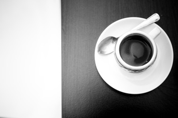 Monochrome picture of top view to a coffee cup with plate and tea spoon on dark table and copy space on left. Espresso geometrical concept. Coffee-mania, black and white. Isolation with hot beverage