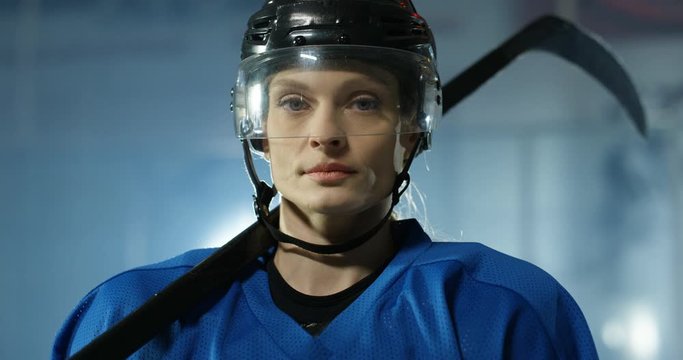 Portrait shot of serious beautiful young Caucasian female hockey player in helmet and with club looking at camera on ice arena. Close up of face of attractive sportswoman in casque with stick.