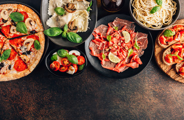 Full table of italian meals on plates Pizza, pasta, ravioli, carpaccio. caprese salad and tomato bruschetta on black background.
