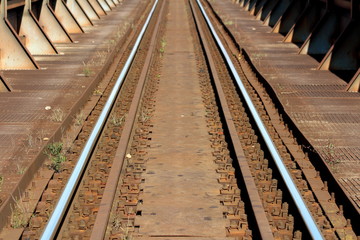 Fototapeta premium Shining metal railway tracks mounted with rows of strong industrial size bolts to old rusted railway bridge leading to railway station mixed with patches of grass growing between cracks
