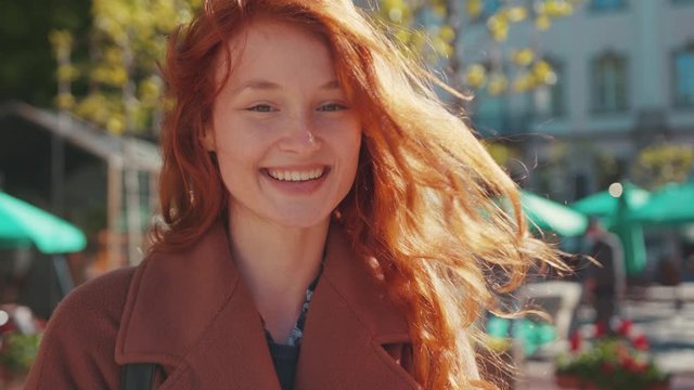 Outdoor portrait of appealing shy cute ginger girl in coat holding smartphone smiling cheerful with much joy. Young people. Fashion. Urban lifestyle.