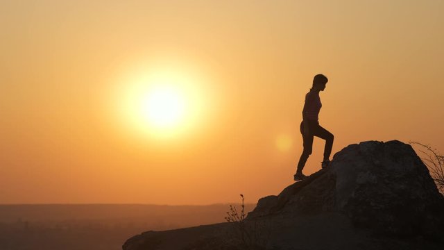 Silhouette of a woman hiker climbing alone on big stone at sunset in mountains. Female tourist raising her hands up on high rock in evening nature. Tourism, traveling and healthy lifestyle concept.